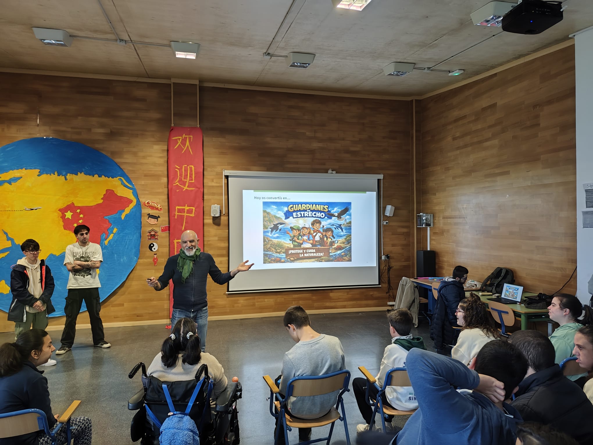 Teacher presenting to a group of seated students in a classroom with a large globe mural and a projector screen showing a poster poster about nature conservation.