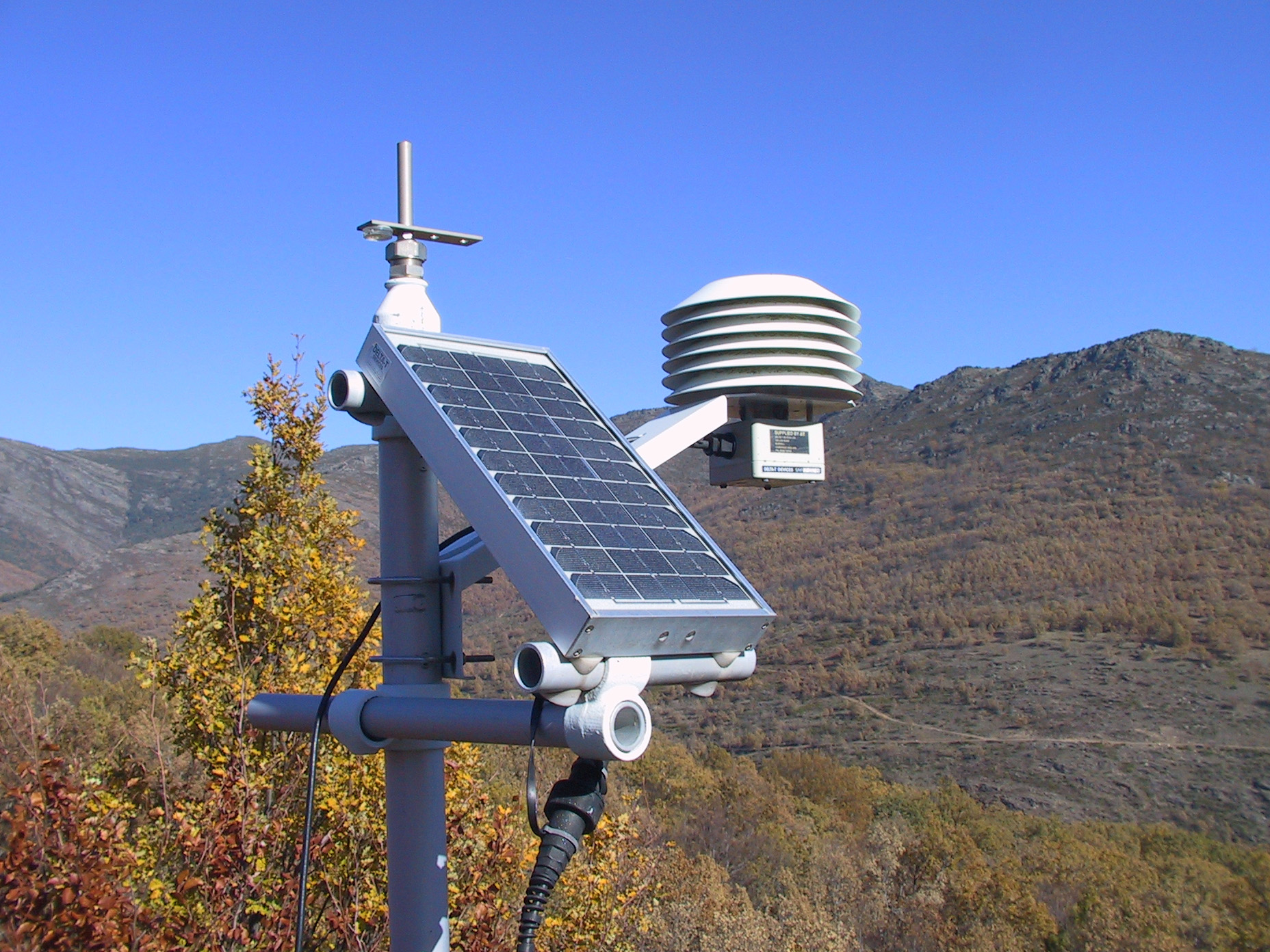 Outdoor weather station with solar panel and vented sensor against a mountain landscape.