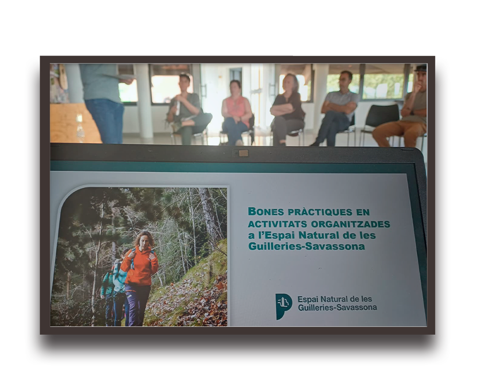 Laptop screen showing a presentation slide about organized activities at Espai Natural de les Guilleries-Savassona; people sit in a room in the background.