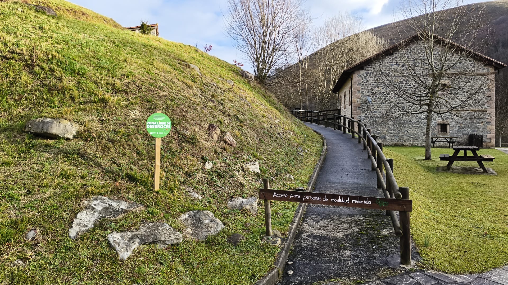 Gravel path with a wooden railing runs alongside a stone building; a green sign on the hill reads 'Zona libre de desbroces' and a picnic area sits nearby, indicating disabled-accessible access.