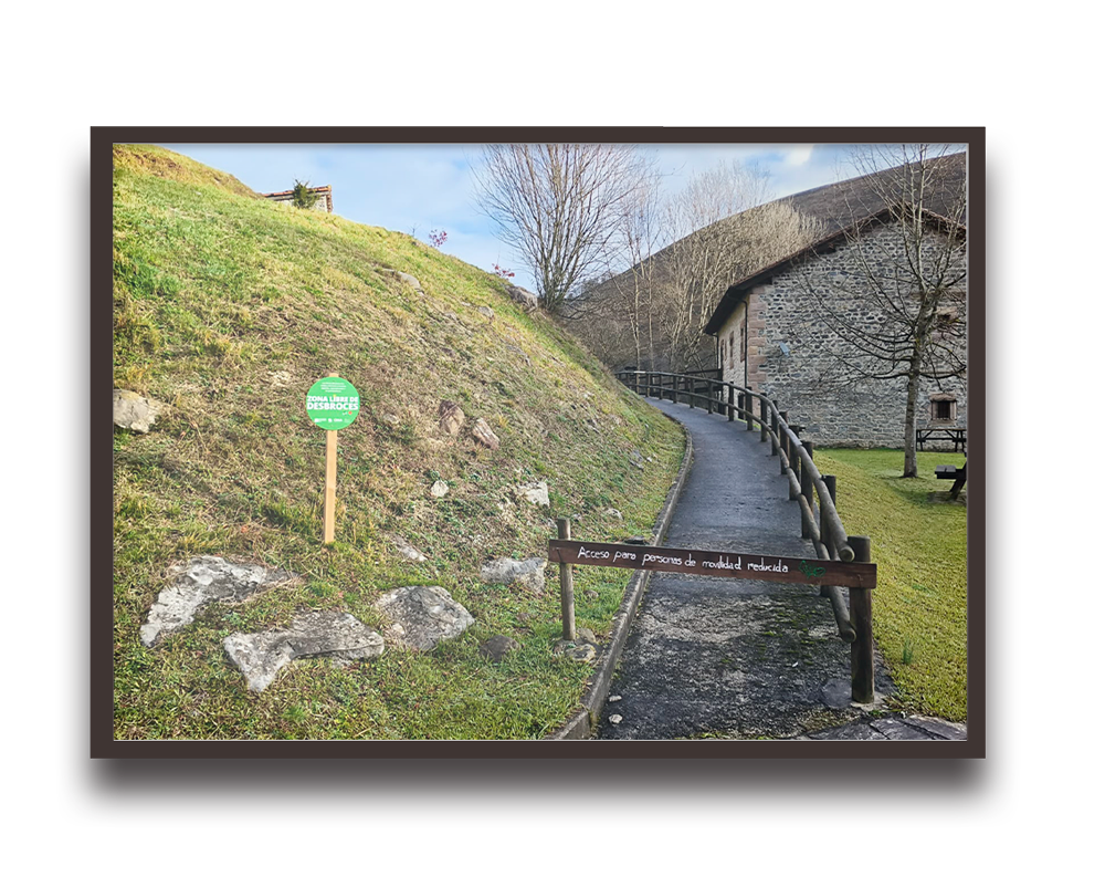 Paved pedestrian path with a wooden barrier and railing leading to a stone building, sign indicates accessibility for people with reduced mobility on the gate top.