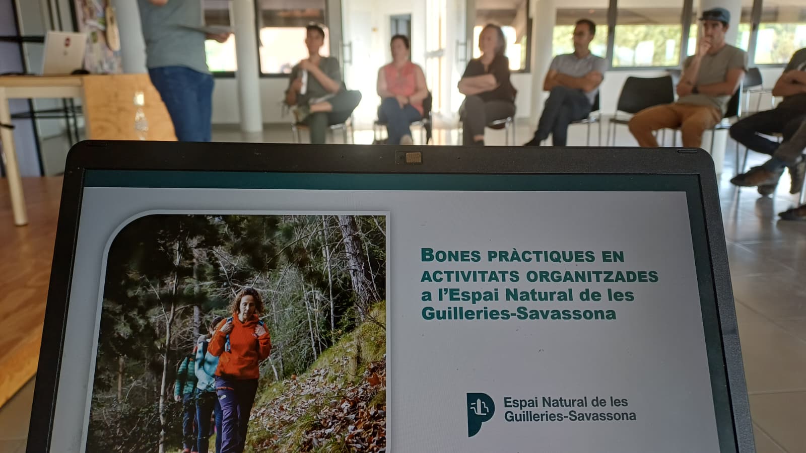 Laptop screen displaying a presentation on best practices for activities at Espai Natural de les Guilleries-Savassona; a group sits in a circle in a bright room behind.