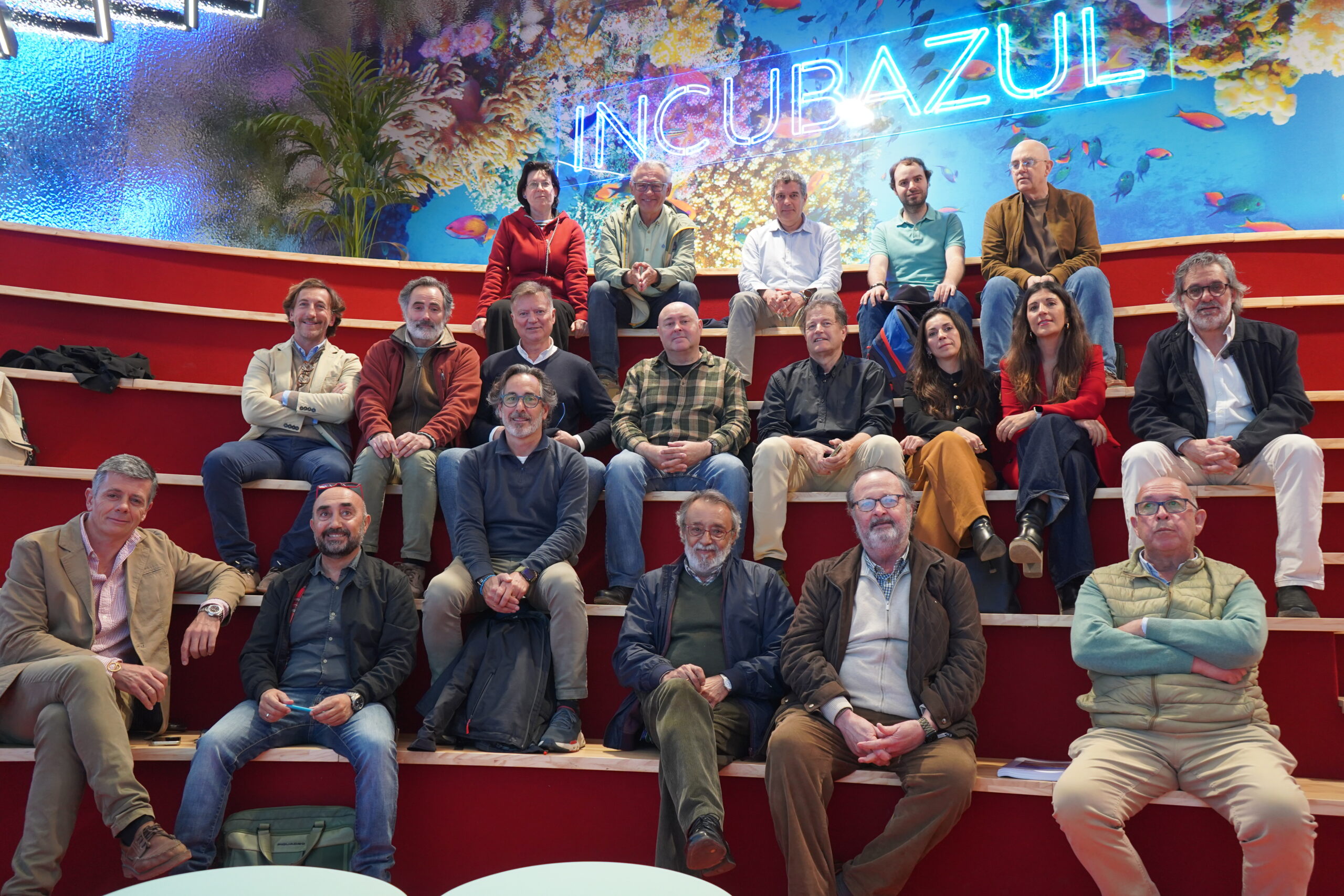 Group of adults seated on red bleachers in front of a bright coral reef mural with a blue neon sign behind them.