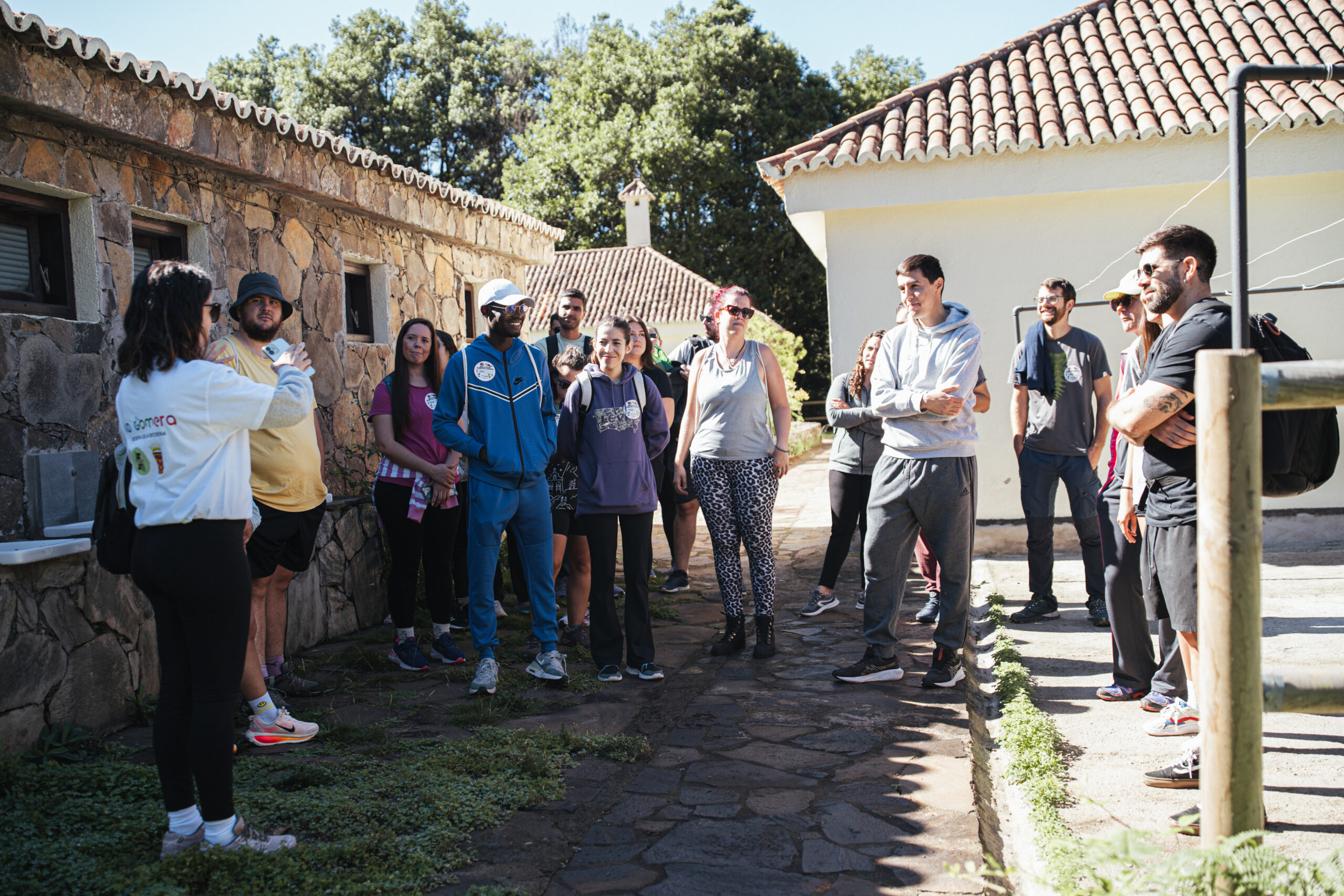 Group of people with backpacks listening to a guide in a sunny stone courtyard with tiled roofs and stone walls north of a trail.