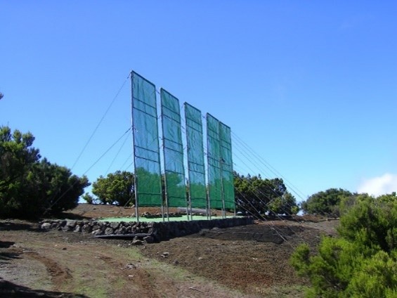 Three tall green mesh panels on metal frames anchored by guy wires, set on a stone retaining wall against a clear blue sky.
