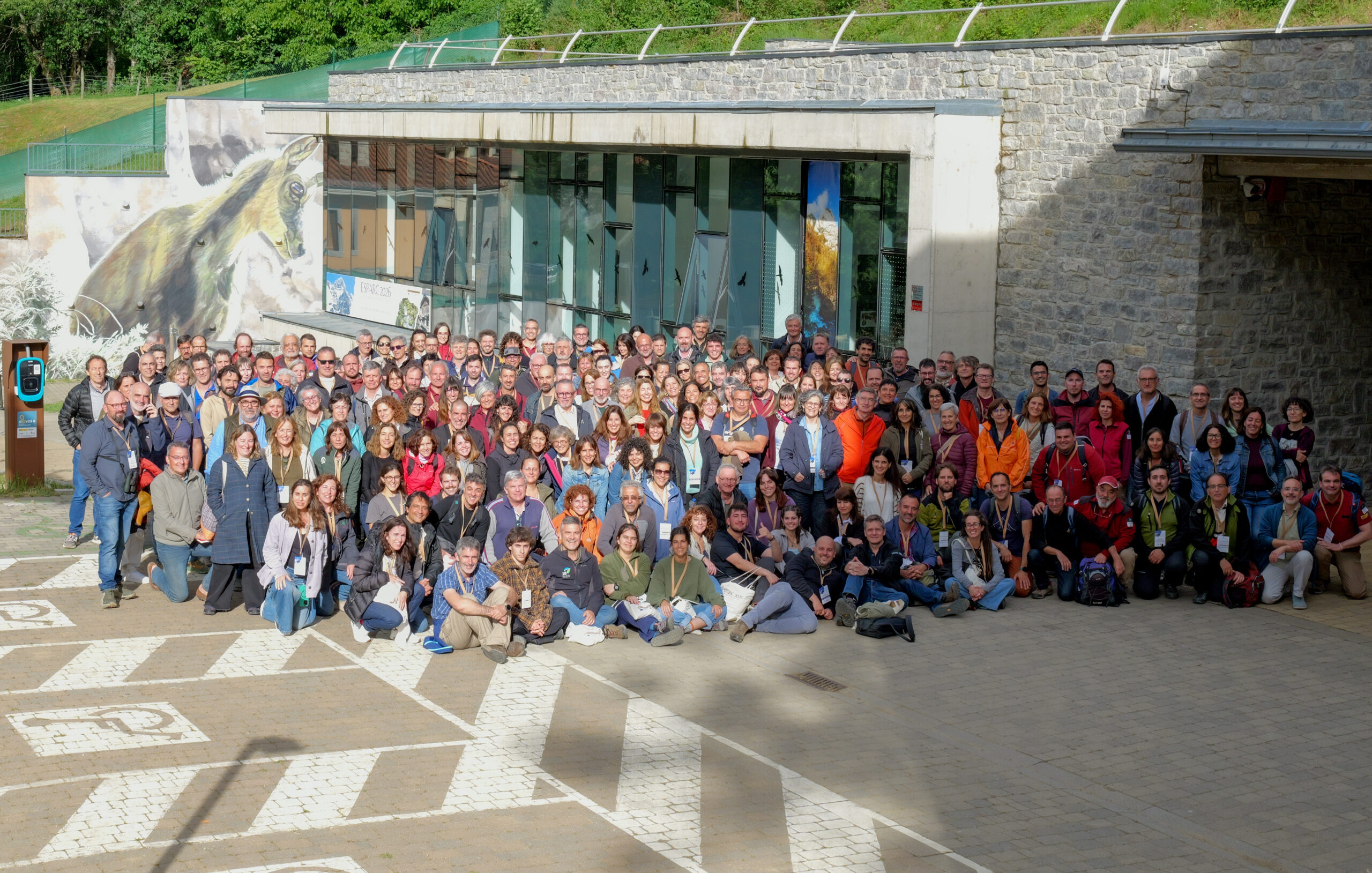 Large group of people posing for a group photo outdoors in front of a glass-fronted building with a horse mural, stone wall, and green hill in the background.