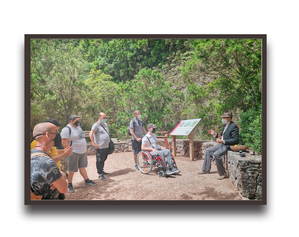 Group of people wearing masks listening to a man in a suit giving an outdoor talk near an information board in a green park.