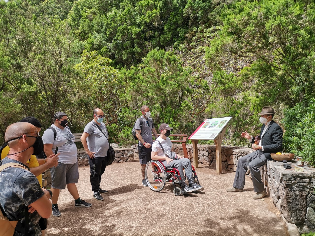 Tour guide in a suit and hat explains something to a small group outdoors; a man in a wheelchair sits nearby as masked visitors listen.