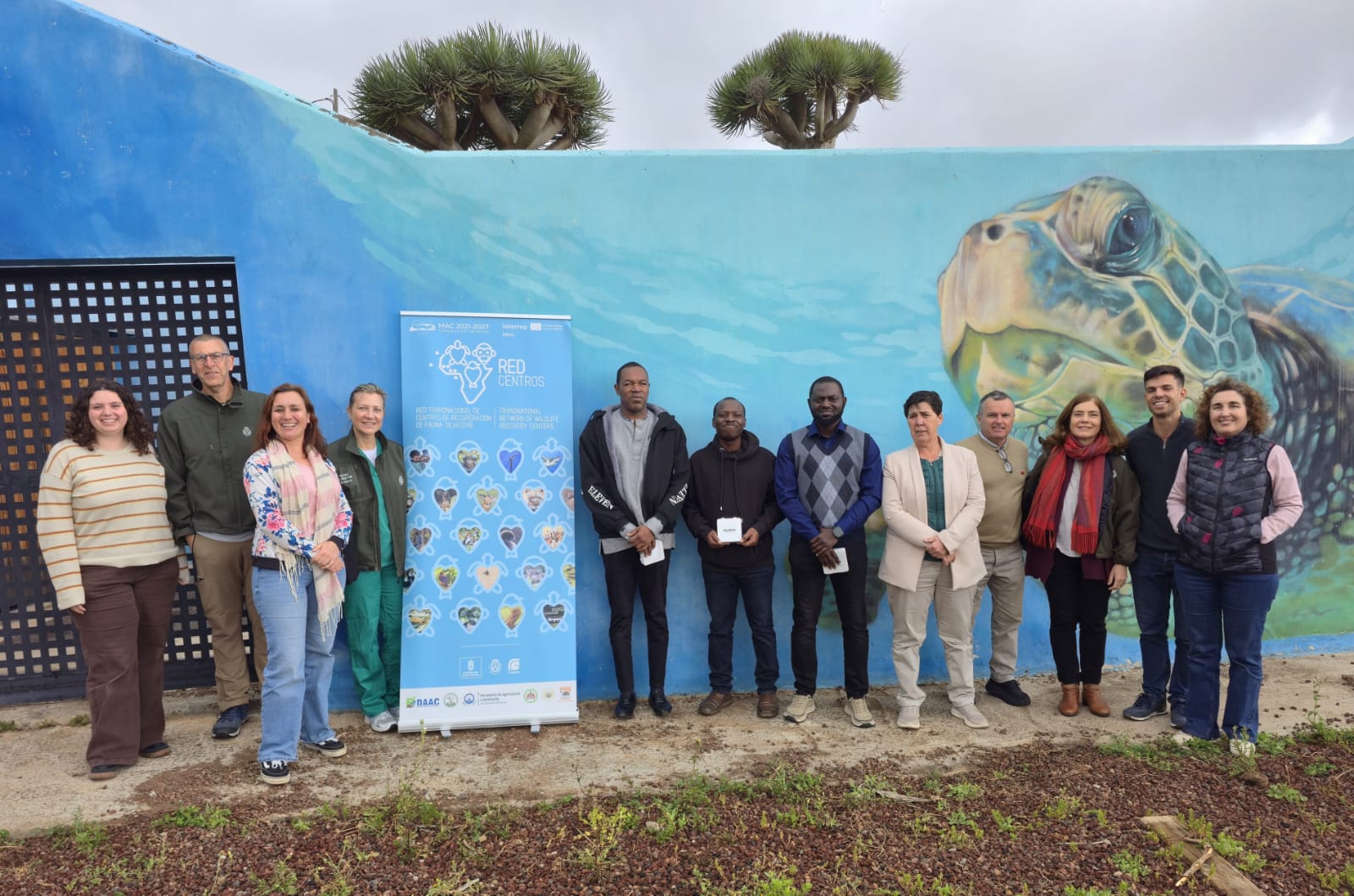 Group of adults posing in front of a blue mural featuring a sea turtle, with a standing banner about Red Centros project nearby.