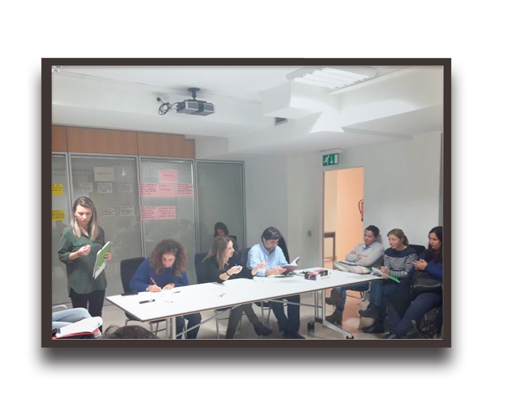 Team meeting in a conference room with a presenter standing at the left while others work at a long table with notes on the wall behind them