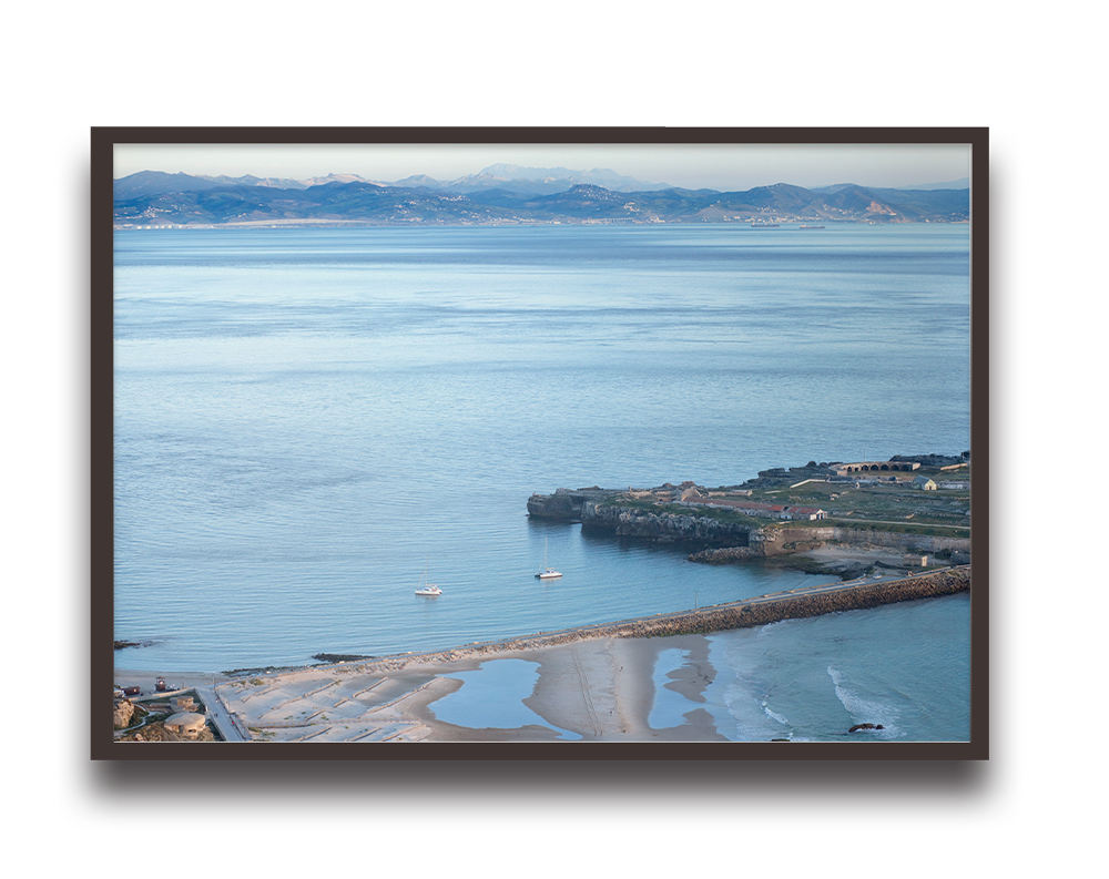 Coastal scene with calm blue waters, small boats, and distant mountains framed by a dark photo border on a wall.