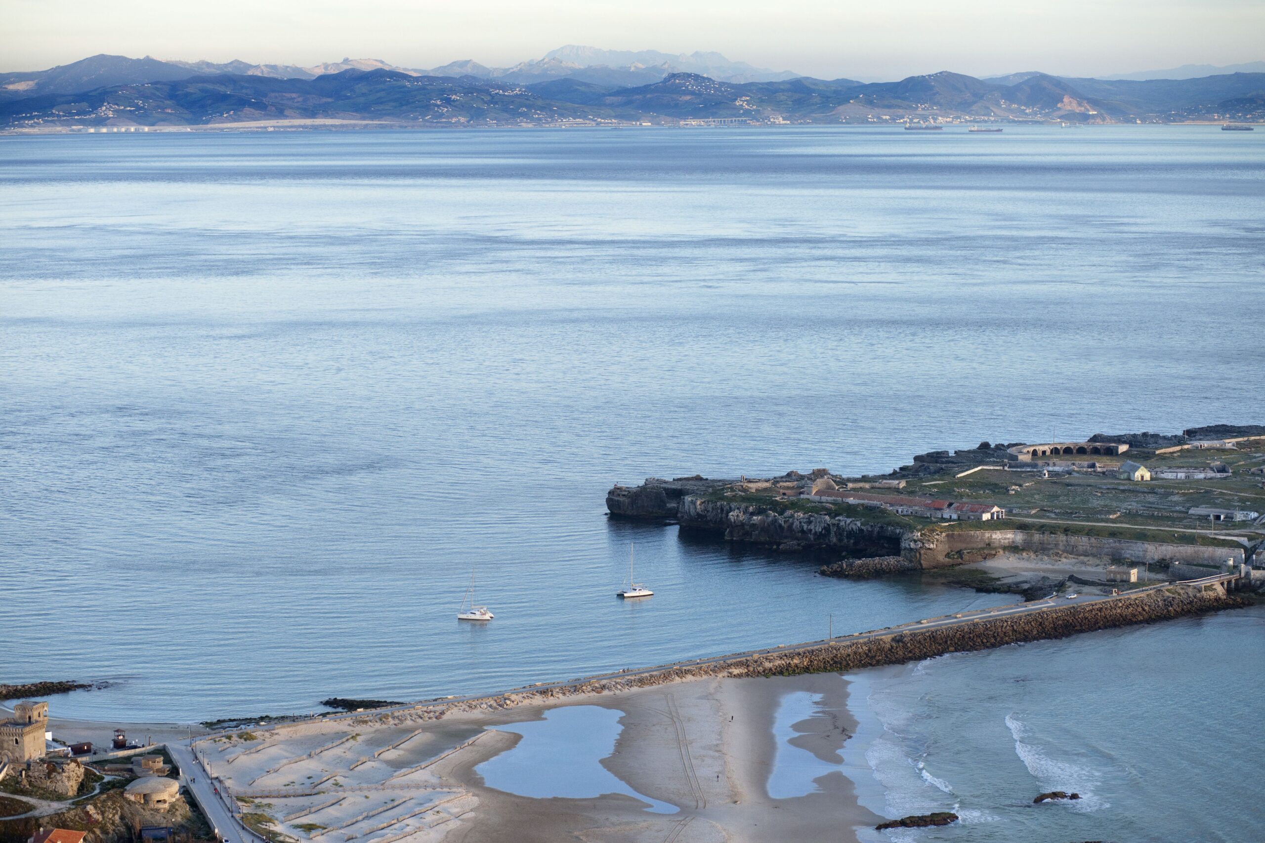 Coastal town along a calm blue bay with distant mountains and small sailboats on the water.