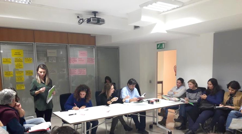 Group of adults seated around a conference table as a woman presents notes standing at the head of the table