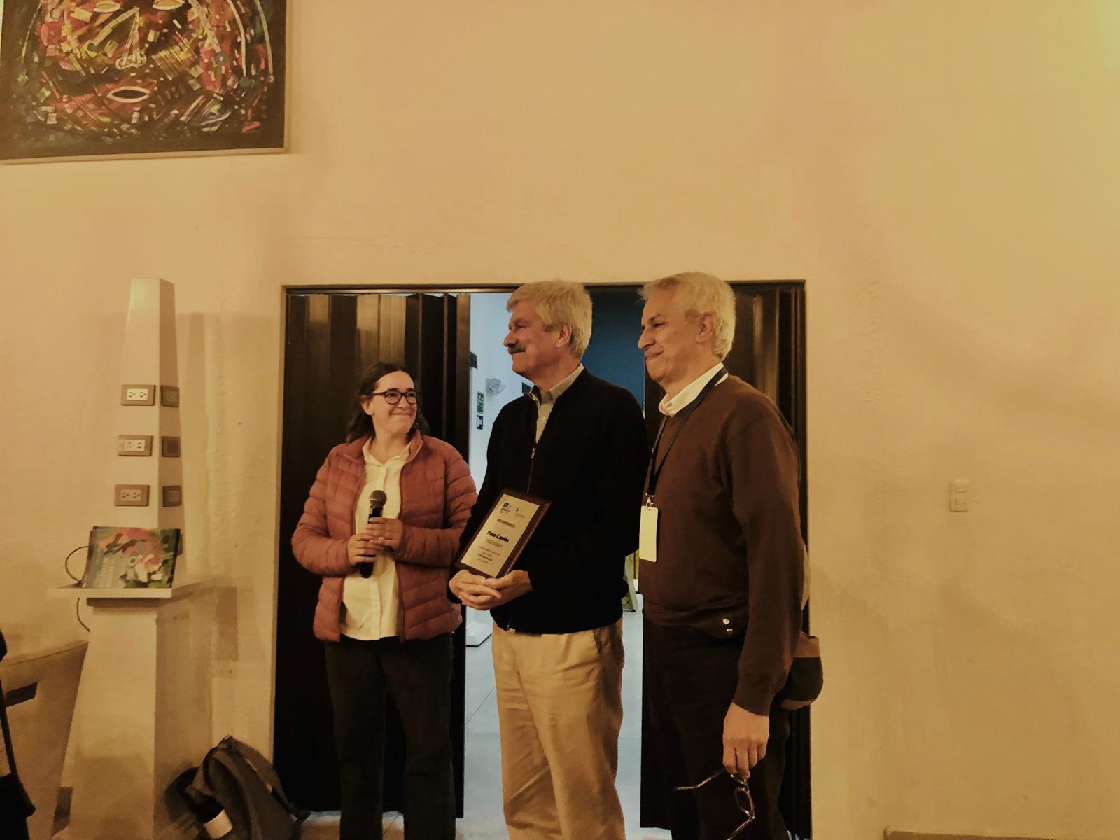 Three adults stand in a doorway at an event: woman with a microphone on the left, man in the middle holding a plaque, and another man on the right wearing a badge.