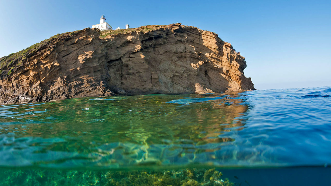 Rocky coastal cliff with a white lighthouse on top, blue sky and clear turquoise water below the cliff.