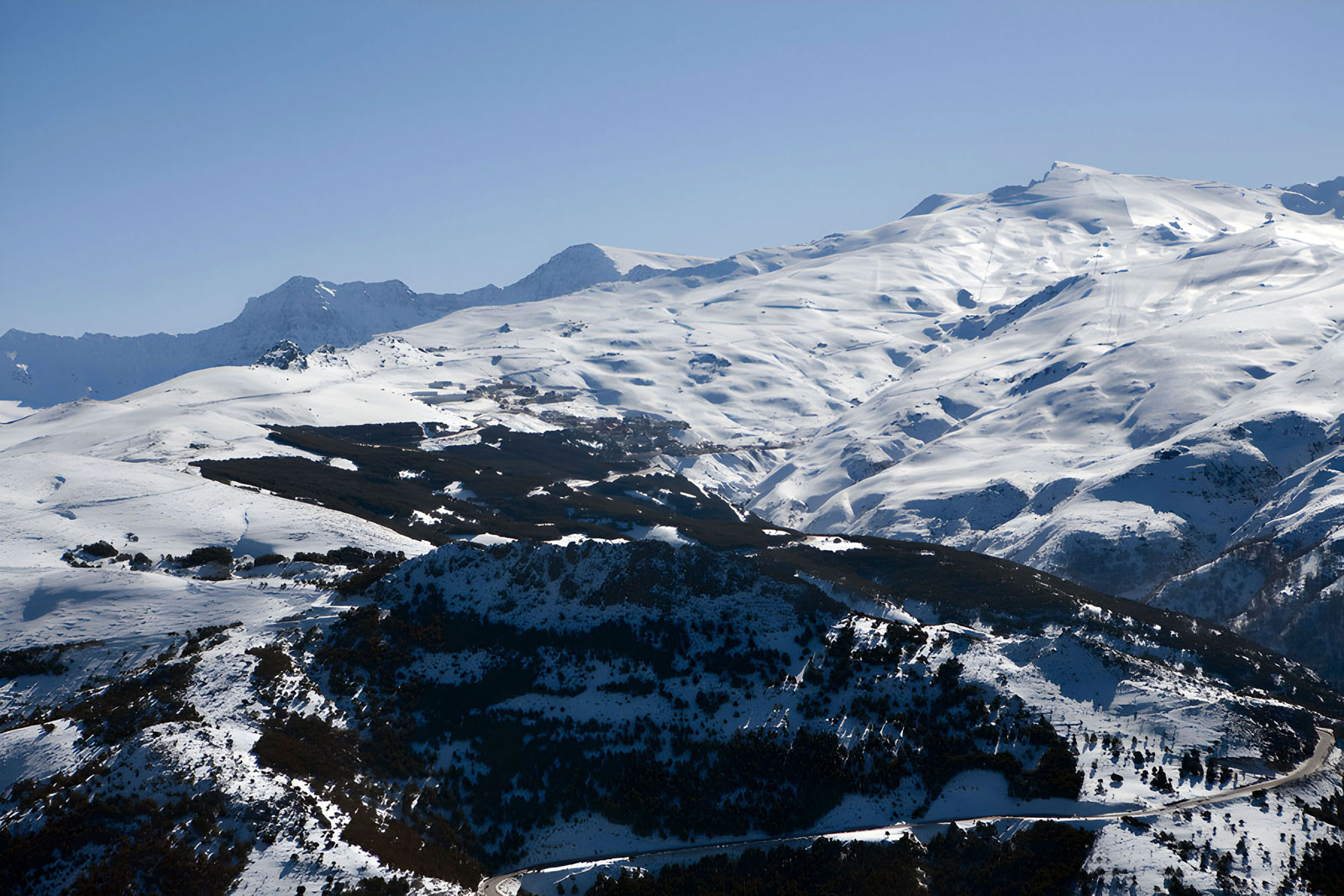 Snow-covered alpine mountains under a clear blue sky, with dark rocky ridges in the foreground.