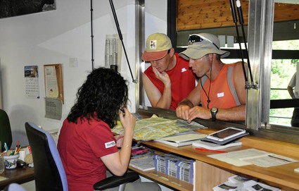 Information desk: a woman in red helps two visitors on the other side of a glass partition with maps and papers on the counter.