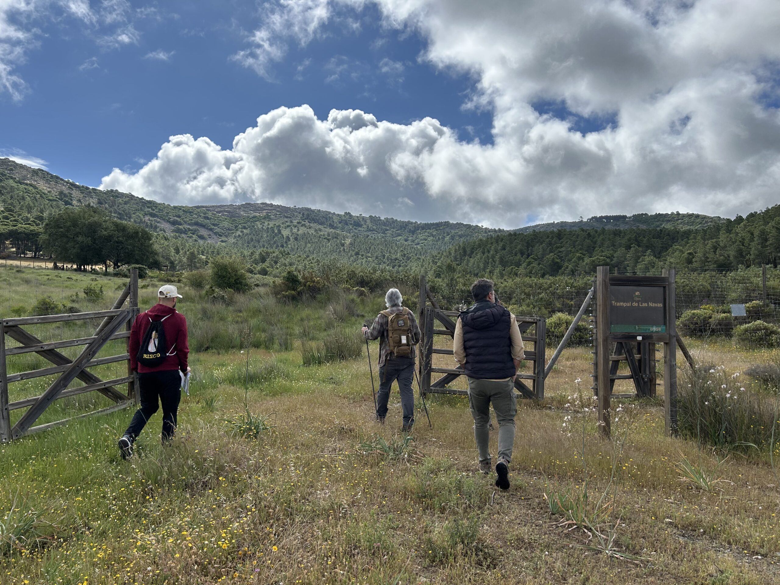 Hikers approach a wooden gate across a grassy meadow with hills and a cloudy blue sky in the background.